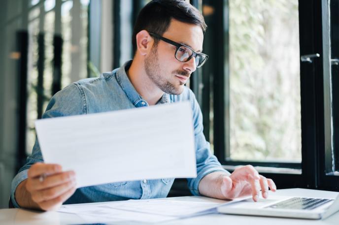 An image of a man on a laptop with a piece of paper in his right hand